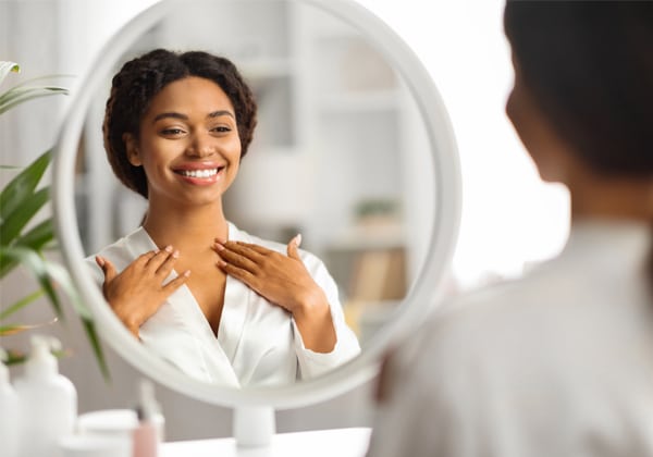 Woman applying skincare product while looking in bathroom mirror, smiling.