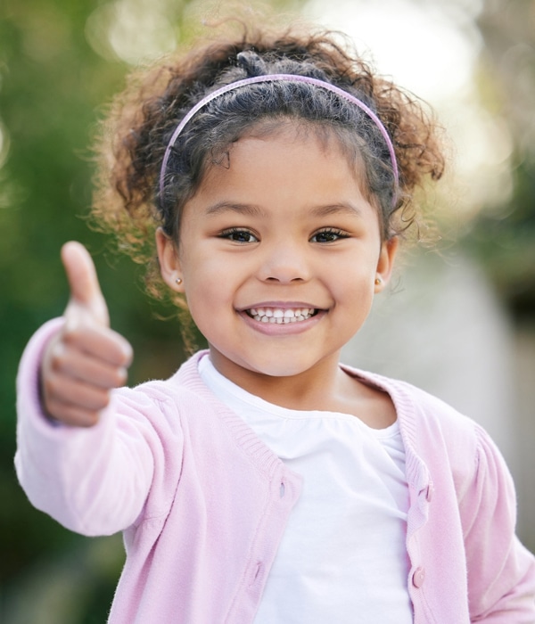 Young child smiling and giving thumbs up outdoors in pink cardigan.