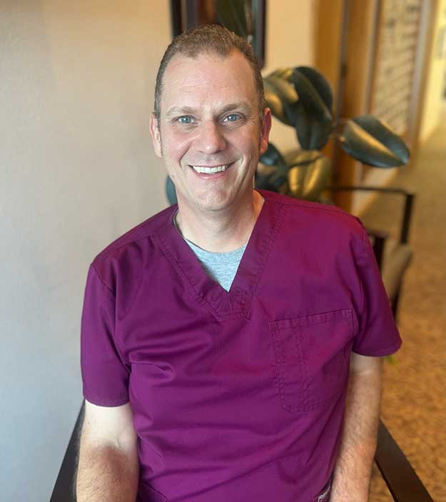 Male dental professional in purple scrubs smiling in dental office interior.