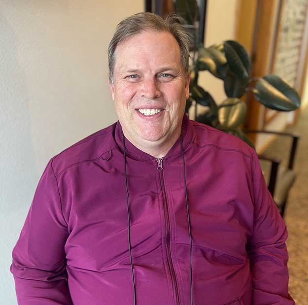 Man in purple shirt smiling indoors with plant background, dental office setting.