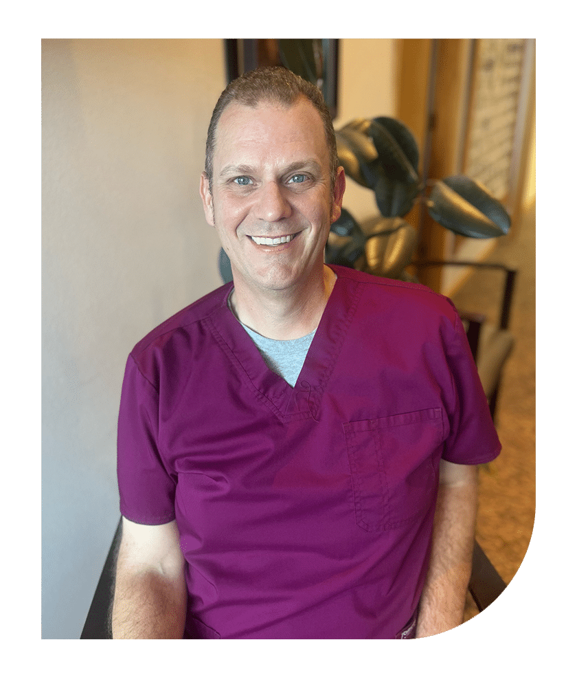Smiling male dentist in purple scrubs inside dental office treatment room.